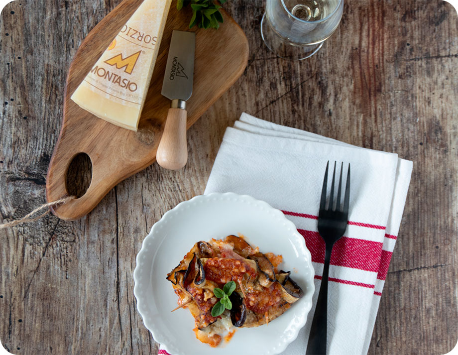 piatto di parmigiana a tavola con a fianco uno spicchio di forma di formaggio montasio su tagliere in legno